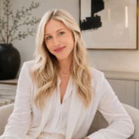 Smiling blonde woman in a white blazer in a modern office with minimalist decor.