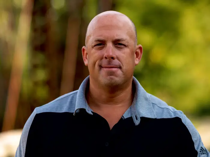 Smiling bald man in a grey and black polo shirt against blurred green foliage.