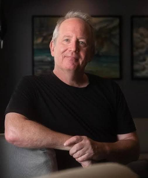 Middle-aged man with gray hair wearing a black t-shirt, smiling in front of framed paintings.
