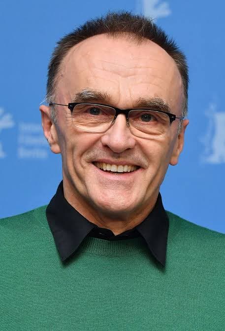 Danny Boyle smiling, wearing glasses and a green sweater over a black collared shirt.