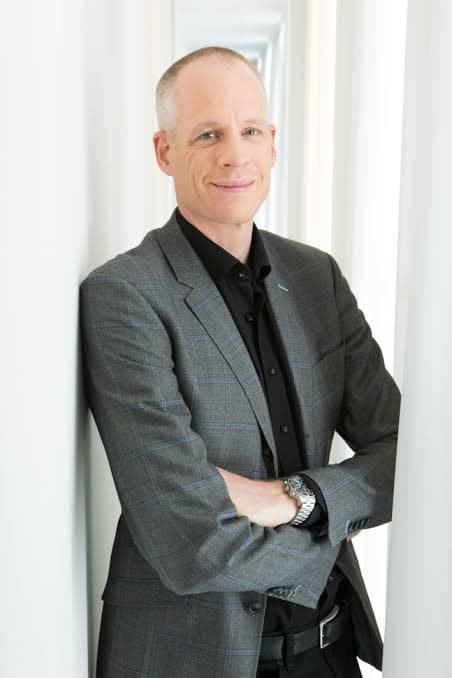 Smiling man in a grey plaid blazer and black shirt leaning against a white wall.