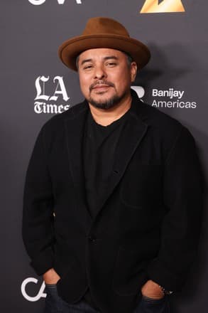 Jacob Vargas wearing a brown fedora and black blazer, posing against a branded backdrop.